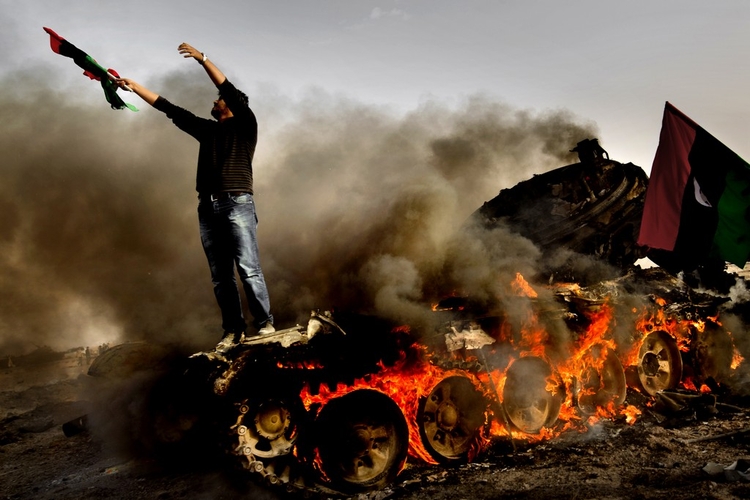 Mężczyzna stoi na płonącym wraku czołgu, wymachując libijską flagą z okresu poprzedzającego rządy Mu'ammara al-Kaddafiego. Jego gest jest protestem w związku z atakiem wojsk pułkownika na Adżdabiję, miasta położonego na południe od Bengazi. 17 lutego 2011; fot. Mads Nissen, Panos Pictures